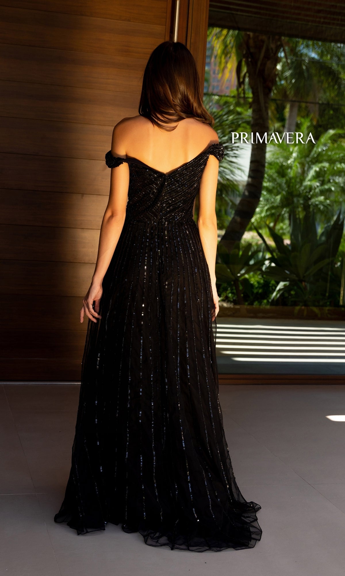A woman in a black, off-the-shoulder Long Prom Dress 13125 by Primavera, featuring sequin details, stands indoors with her back turned. The word